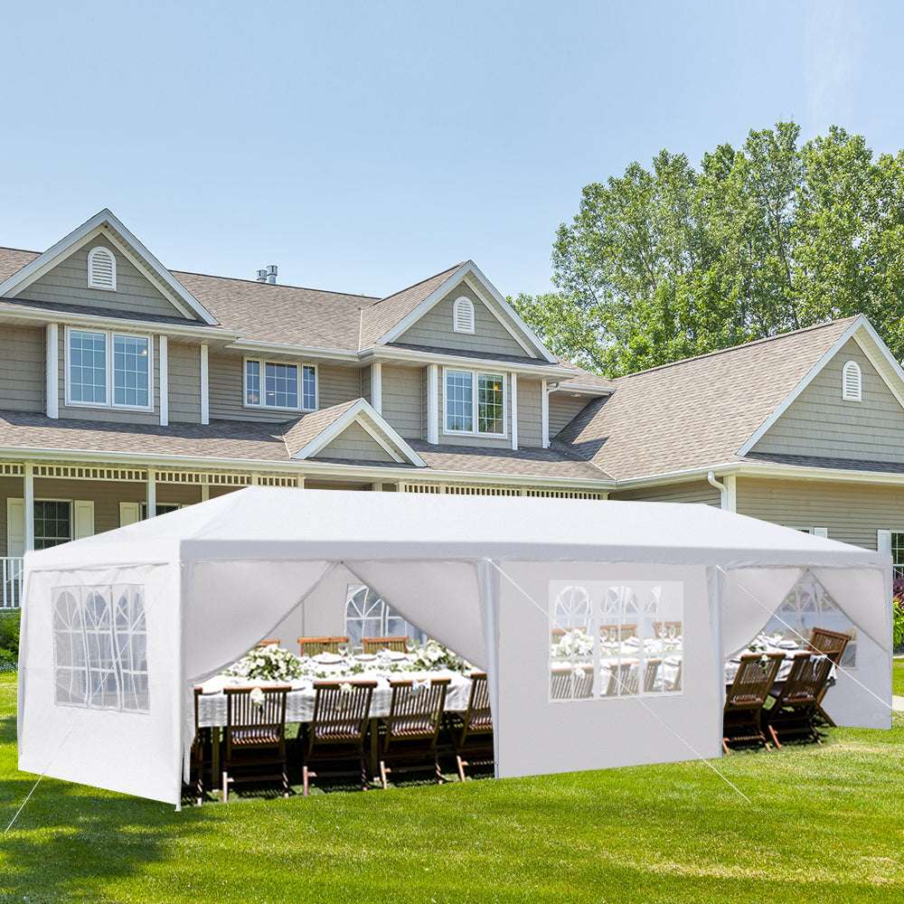 White outdoor canopy tent set up on a grassy lawn in front of a house with tables and chairs underneath.
