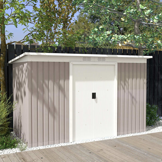Metal garden storage shed in soft taupe with white trim, placed on decking against a black fence and leafy backdrop.