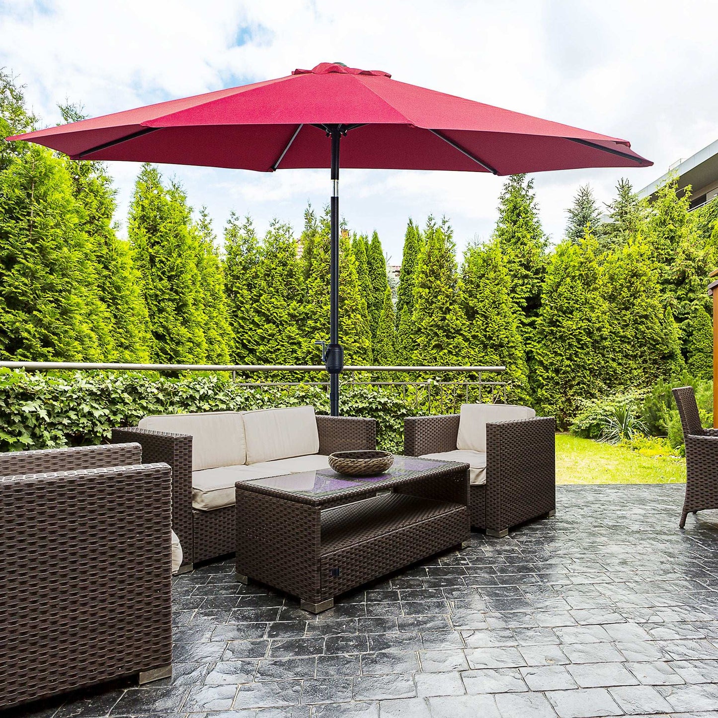 Outdoor patio set with red umbrella, sofa, and coffee table on a stone patio with greenery in the background.