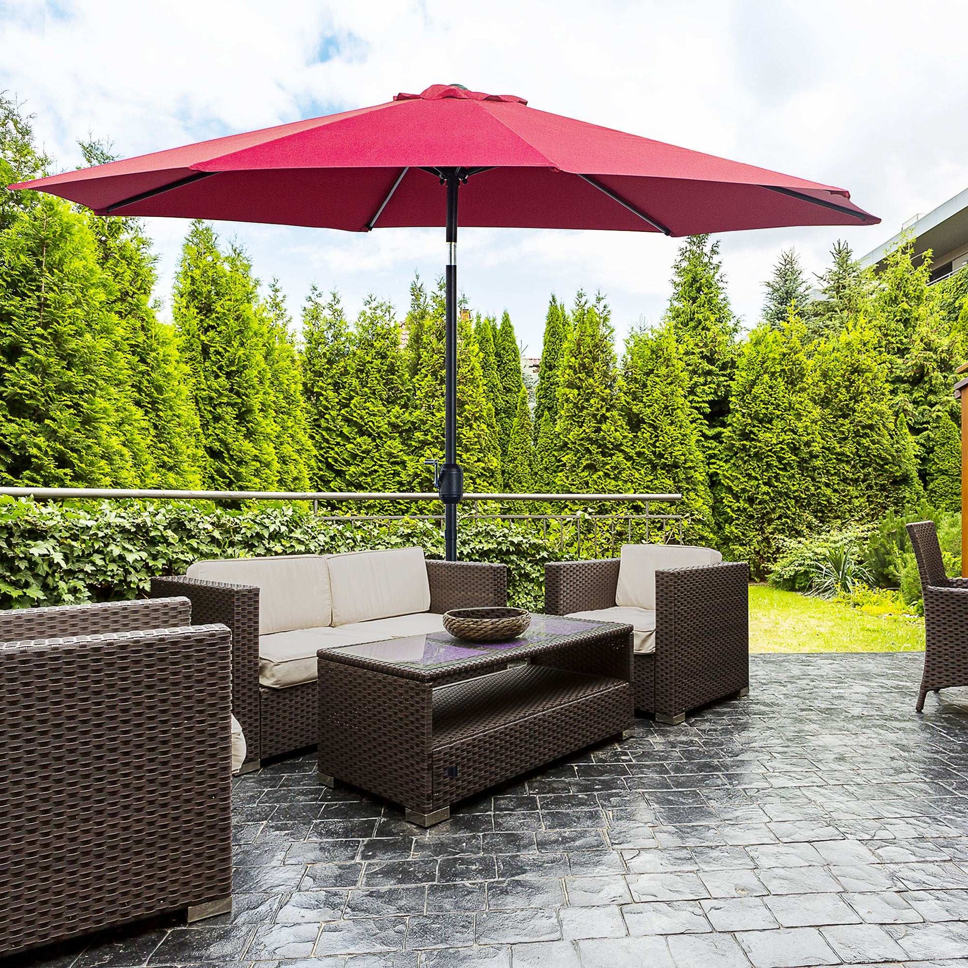 Outdoor patio set with red umbrella, sofa, and coffee table on a stone patio with greenery in the background.