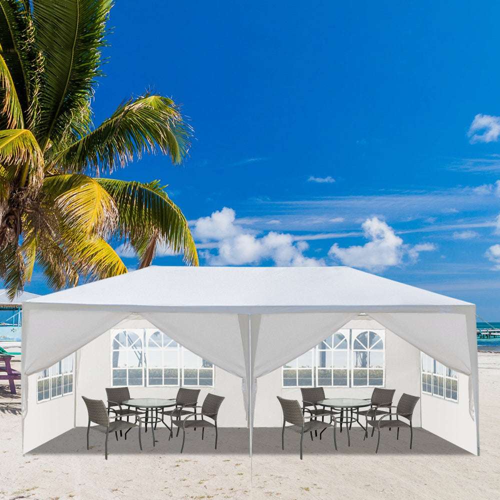 White outdoor canopy with tables and chairs on a beach with palm trees and blue sky.